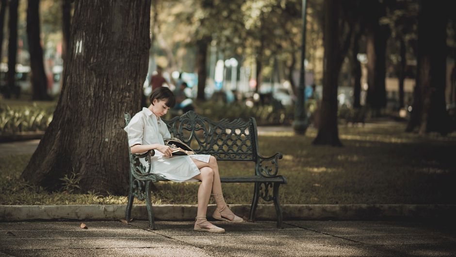 A woman in a white dress enjoys reading a book on a park bench surrounded by nature.